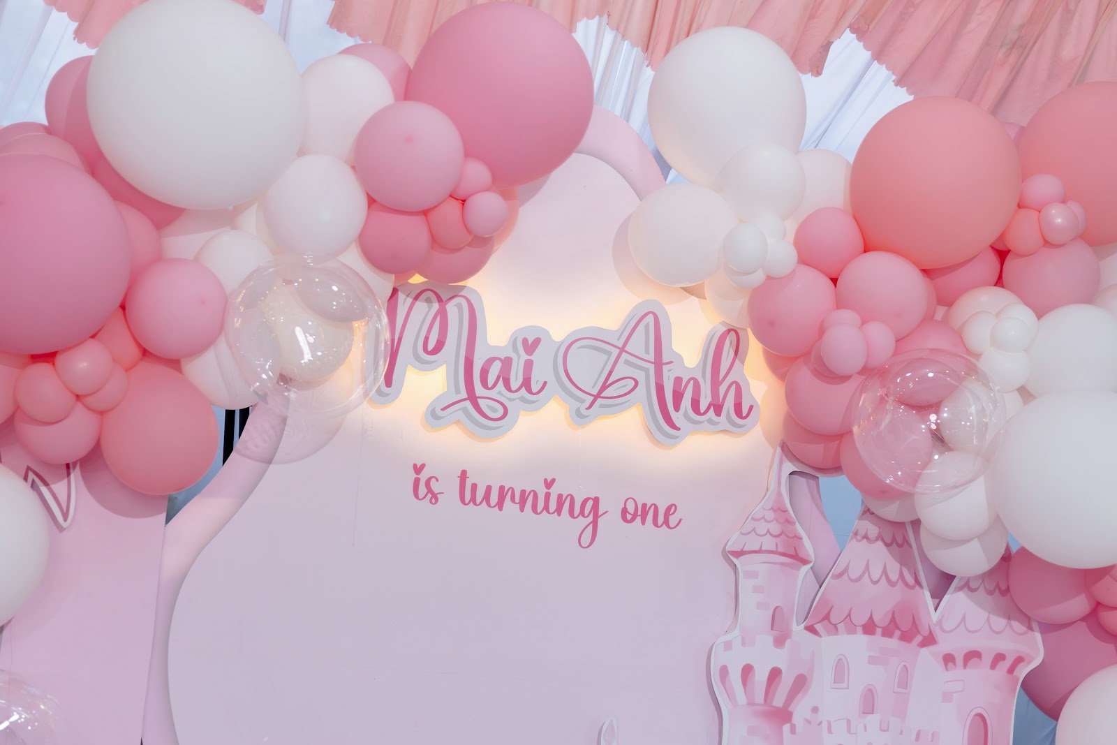 Chữ nổi gắn đèn LED tên bé kèm dòng chữ is turning one trên backdrop thôi nôi tông hồng pastel