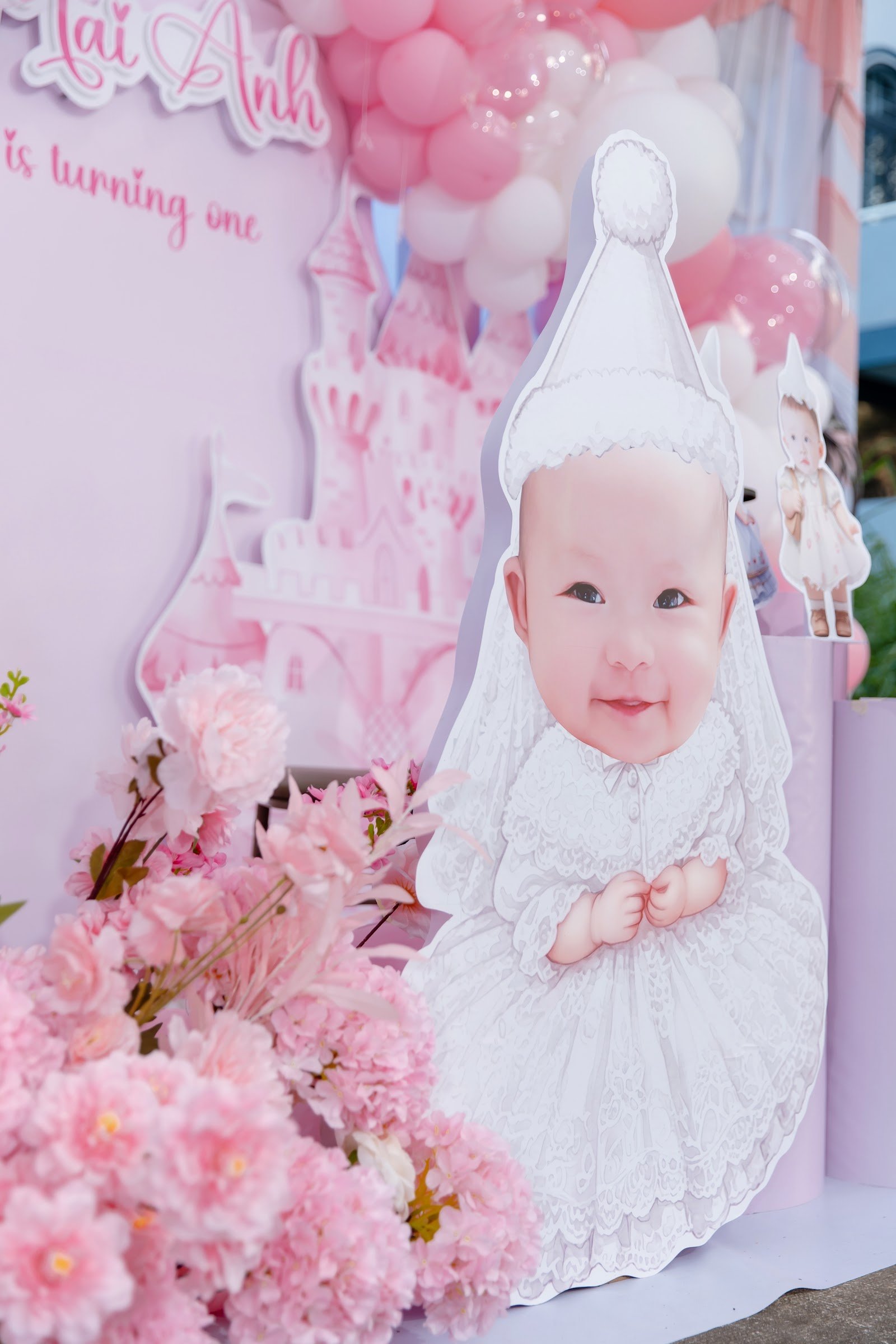 Standee in hình bé gái mặc váy trắng đặt giữa cụm hoa tươi tông hồng tại tiệc thôi nôi